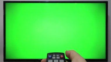 TV kumandasını gösteren ve yeşil ekranlı bir televizyonun önündeki düğmelere basan kişi