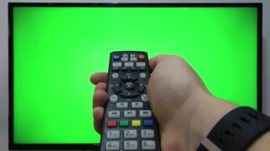 TV kumandasını gösteren ve yeşil ekranlı bir televizyonun önündeki düğmelere basan kişi