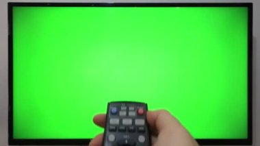 TV kumandasını gösteren ve yeşil ekranlı bir televizyonun önündeki düğmelere basan kişi