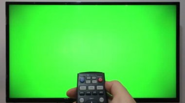 TV kumandasını gösteren ve yeşil ekranlı bir televizyonun önündeki düğmelere basan kişi