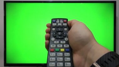 TV kumandasını gösteren ve yeşil ekranlı bir televizyonun önündeki düğmelere basan kişi