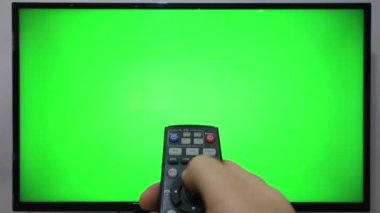 TV kumandasını gösteren ve yeşil ekranlı bir televizyonun önündeki düğmelere basan kişi