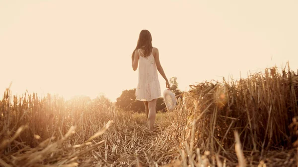 Ragazza felice sulla strada in un campo di grano al tramonto, Ragazza a piedi nudi in un vestito bianco che cammina sul campo di grano Fotografia Stock