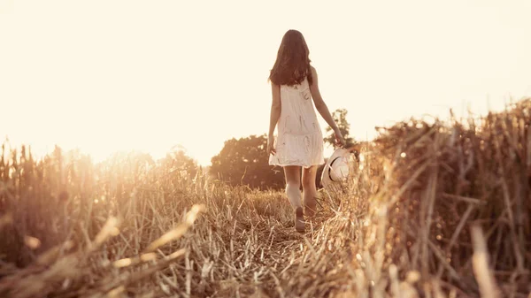 Gelukkig meisje op de weg in een tarweveld bij zonsondergang, meisje in een witte jurk lopen op tarweveld barefoot Stockafbeelding