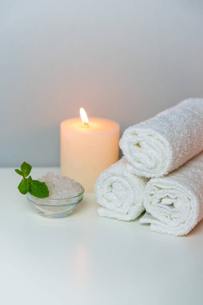 Wellness and SPA concept photo with white towels stack, candle light, sea salt and mint leaf, vertical orientation.