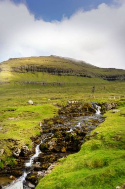 Dağ nehri yokuş aşağı akar, yeşil dağ ve ufukta bulutlu gökyüzü. Eysturoy Adası, Faroe.