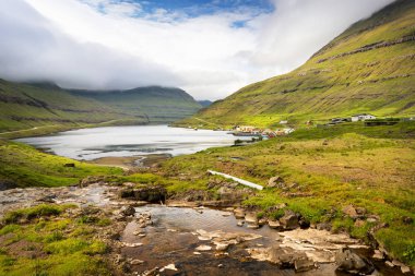 Küçük bir kasaba yakınlarındaki göl aynası ve Faroe Adaları 'ndaki tüylü bulutlarla kaplı yeşil dağlar..
