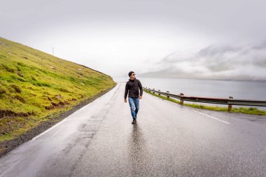 Young traveller walking y empty road after rain, green mountains, fjord and overcast sky on horizon. Faroe Islands, Denmark. clipart