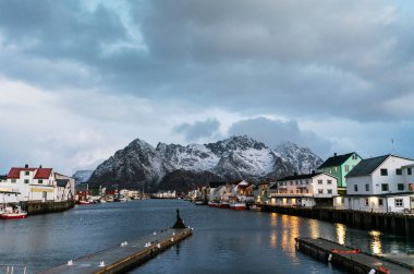 Henningsvaer liman fotoğrafı. Su, dağlar ve ufukta bulutlu gökyüzü yansıyan binalar..