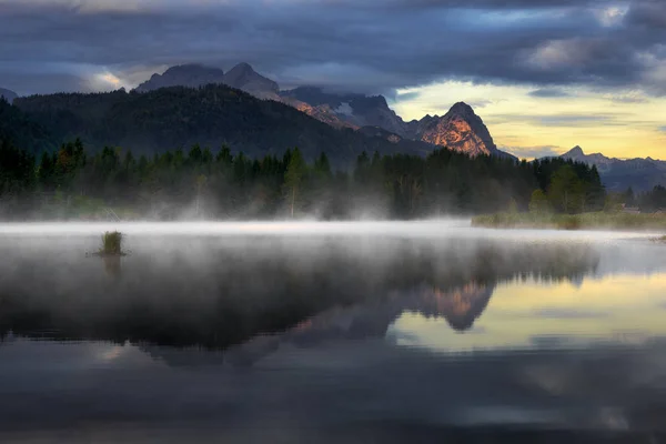 Reflexão da montanha de Wetterstein durante o dia de outono com nevoeiro matinal sobre o lago Geroldsee, Alpes da Baviera, Baviera, Alemanha . — Fotografia de Stock