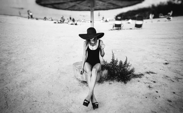 Young pretty girl sitting on the beach in black hat — Stock Photo, Image