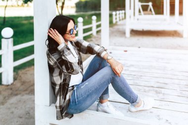 Beyaz ahşap katta oturan ve parkta açık rahatlatıcı güneş gözlüklü genç esmer kız