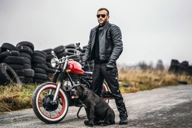 Deri elbiseli bir bisikletçi ve köpeği yolda kırmızı bir motosikletin yanında duruyor. Arka planda çok lastik var.
