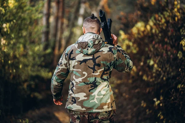 Een soldaat in camouflage uniform met een geweer op zijn schouder lopen in het bos. Achteraanzicht. Airsoft — Stockfoto