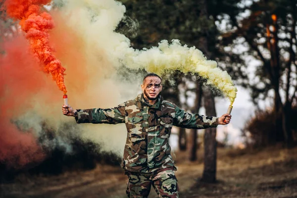 Man in camouflage uniform met zwarte strepen op zijn gezicht in het bos. In haar handen houdt ze rode en witte rookbommen vast. — Stockfoto