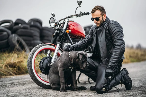 Biker in a leather suit crouched near his dog and red motorcycle on the road. Many tires on the background