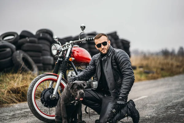 Motorkář v koženém obleku krčil v blízkosti svého psa a červené motocyklu — Stock fotografie