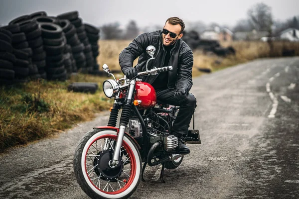 Homem barbudo de óculos de sol e jaqueta de couro sorrindo enquanto sentado em uma motocicleta vermelha na estrada. Atrás dele está uma fila de pneus — Fotografia de Stock