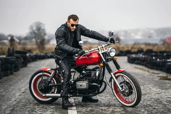 Κόκκινη μοτοσικλέτα με αναβάτη. Ένας άντρας με μαύρο δερμάτινο μπουφάν και παντελόνι στέκεται πλάγια στη μέση του δρόμου. Ελαστικά τοποθετούνται στο παρασκήνιο — Φωτογραφία Αρχείου