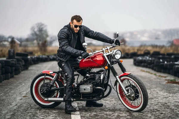 Motocicleta vermelha com piloto. Um homem com uma jaqueta de couro preto e calças fica de lado no meio da estrada. Pneus são colocados no fundo — Fotografia de Stock