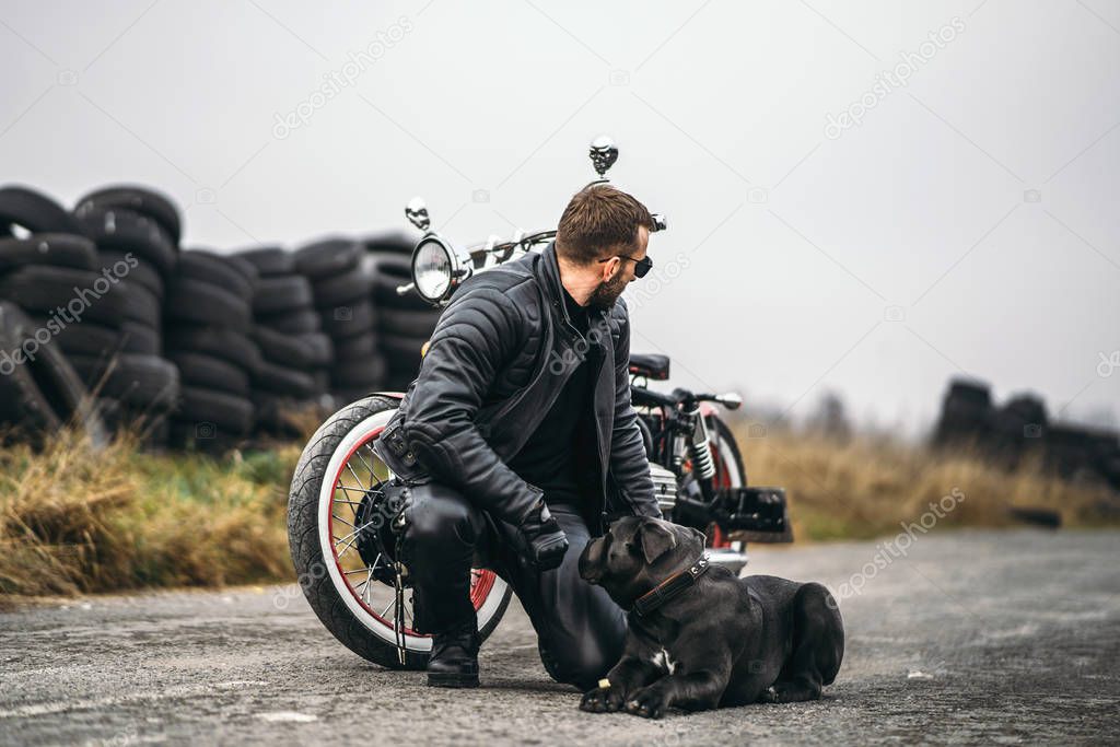 Biker in a leather suit crouched near his dog and red motorcycle on the road. Many tires on the background