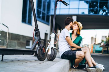Modern cam bir binanın yanında oturan ve öpüşen kadın ve erkeğin bulanık portresi. Elektrikli scooterlarıyla. Ön planda elektrikli scooterları var.