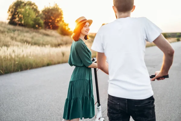 Happy couple walking on scooters. A young woman in dress and hat on sunset background. In the foreground, a man turns his back on the lens — Stockfoto