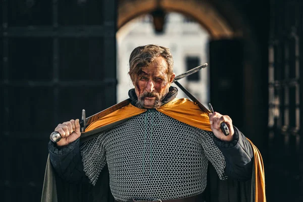 Retrato de um guerreiro sênior medieval em armadura após uma batalha com sangue em seu rosto. O guerreiro tem duas espadas nas mãos — Fotografia de Stock