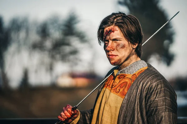 Retrato de um jovem guerreiro medieval de armadura com espada no ombro. No processo de batalha, com feridas no rosto — Fotografia de Stock