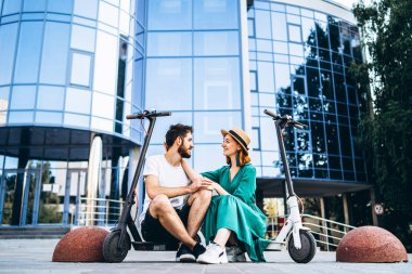Genç ve sevgi dolu çift elektrikli scooterlarıyla modern cam bir binanın yanında oturuyorlar. Büyük şehirde yürür