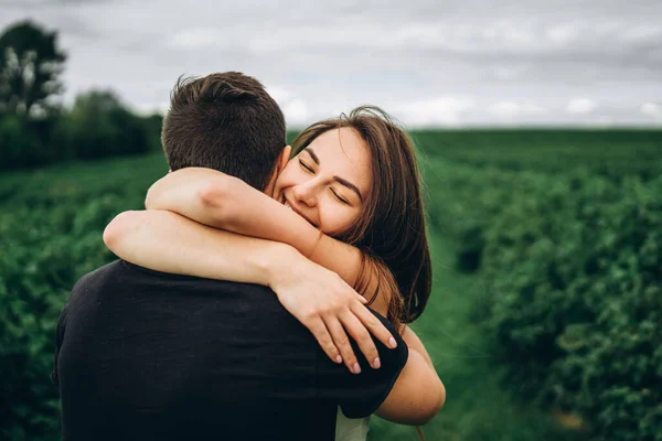 Nettes junges Mädchen mit langen Haaren umarmt ihren Liebhaber lächelnd und mit geschlossenen Augen. junges Paar spaziert in einem Feld in der Natur — Stockfoto