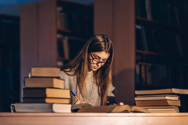 Junger Student mit Brille bereitet sich auf die Prüfung vor. Mädchen am Abend sitzt an einem Tisch in der Bibliothek mit einem Stapel Bücher — Stockfoto