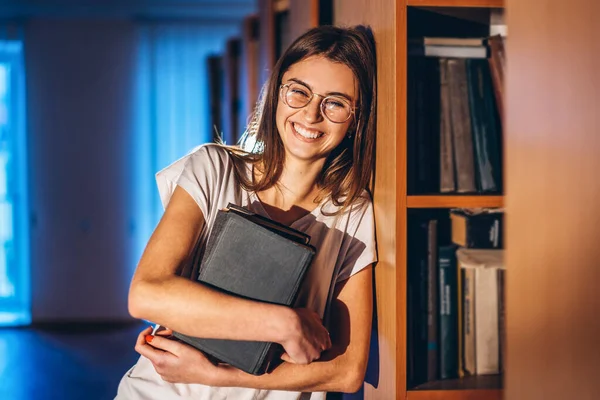 Junge Studentin mit Brille in Bibliothek steht neben Bücherregal — Stockfoto