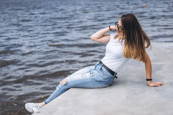 年轻的女人 留着长发 戴着时髦的眼镜 躺在湖边的混凝土岸边 穿着牛仔裤和T恤的女孩面带微笑地看着远方 — 图库照片