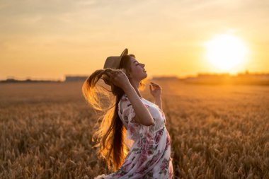 Uzun kıvırcık saçlı, inanılmaz bir genç kadın. Gün batımında buğday tarlasında poz veren ve saçları geriye doğru eğilmiş bir kadın..