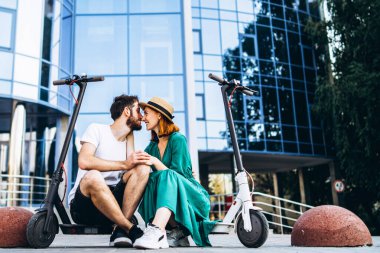 Genç ve sevgi dolu çift elektrikli scooterlarıyla modern cam bir binanın yanında oturuyorlar. Büyük şehirde yürür.