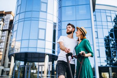 Elektrikli scooterlı genç ve romantik bir çift şehirde yürüyor. Şapkalı genç kadın ve erkek yürümekten zevk alıyor..