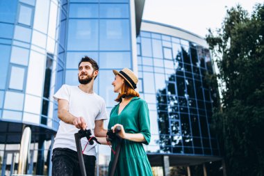 Elektrikli scooterlı genç ve romantik bir çift şehirde yürüyor. Şapkalı genç kadın ve erkek yürümekten zevk alıyor..