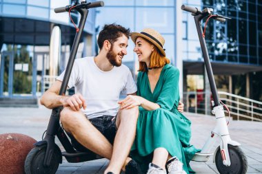 İki çekici insan cam binanın yanında elektro scooterlarıyla takılıyor. Erkek ve kadın tatilin tadını çıkarın..
