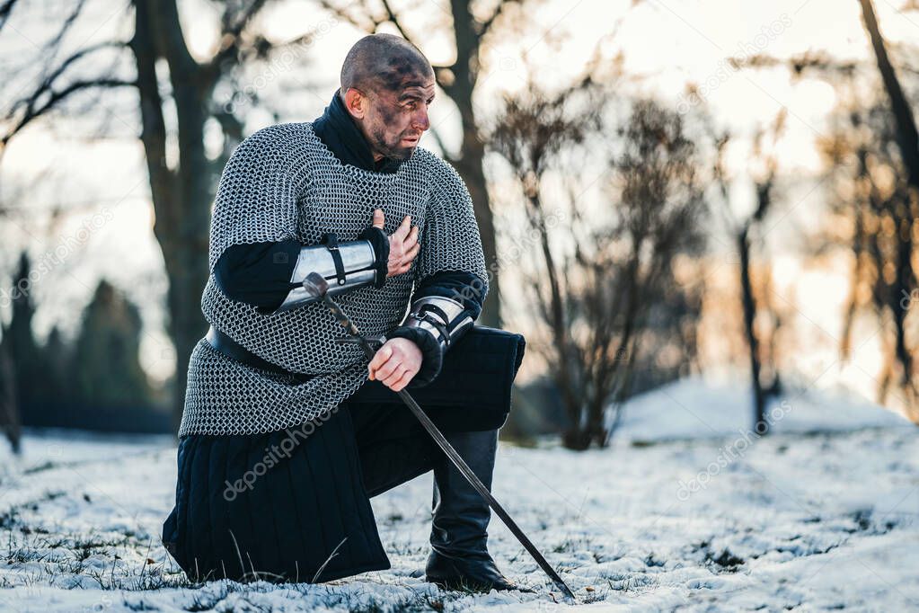 A medieval warrior in chain mail armor kneeling with his sword in his hand and dirty face after the battle. Background of forest and snow.