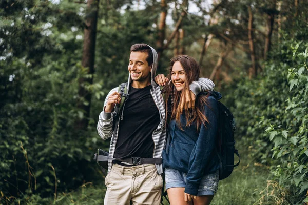 背中をバックパックで笑顔と森の中を歩く若いカップルは 散歩をお楽しみください — ストック写真