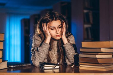 Akşamları kütüphanede koluna yaslanmış kitaplarla oturan yorgun öğrenci kız. Sınavlar için eğitim ve hazırlık.