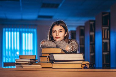 Mutlu gülümseyen bayan öğrenci sınav hazırlığını tamamladı. Kız gece kütüphanede oturan bir yığın kitaba yaslandı..