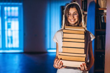 Kütüphanede gözlüklü genç bir kız gülümsüyor ve bir yığın kitap taşıyor. Sınav hazırlığı.