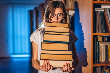 Kütüphanede gözlüklü genç bir kız gülümsüyor ve bir yığın kitap taşıyor. Sınav hazırlığı.