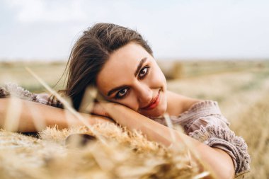 Gözleri kapalı, gülümseyen güzel bir kadının yakın plan portresi. Esmer, bir saman balyasına yaslandı. Arka planda bir buğday tarlası.