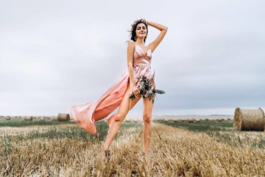 Saman balyalarının yanındaki buğday tarlasında pembe saten elbiseli genç esmer. Kafasında çelenk olan bir kadın elinde bir buket kır çiçeği tutar..