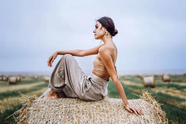 Barefoot brunette in linen pants and bare shoulders sitting on a hay bales in warm autumn day. Behind her is a wheat field.