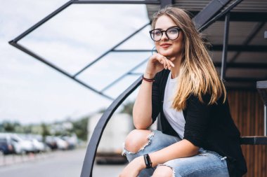 Uzun saçlı ve gözlüklü güzel kız dikey tahtalı evin ahşap arka planında metal merdivenlerde oturuyor. Kadın gülümsüyor ve kameraya bakıyor..