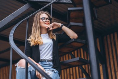 Uzun saçlı ve gözlüklü güzel kız dikey tahtalı evin ahşap arka planında metal merdivenlerde oturuyor. Kadın gülümsüyor ve kameraya bakıyor..